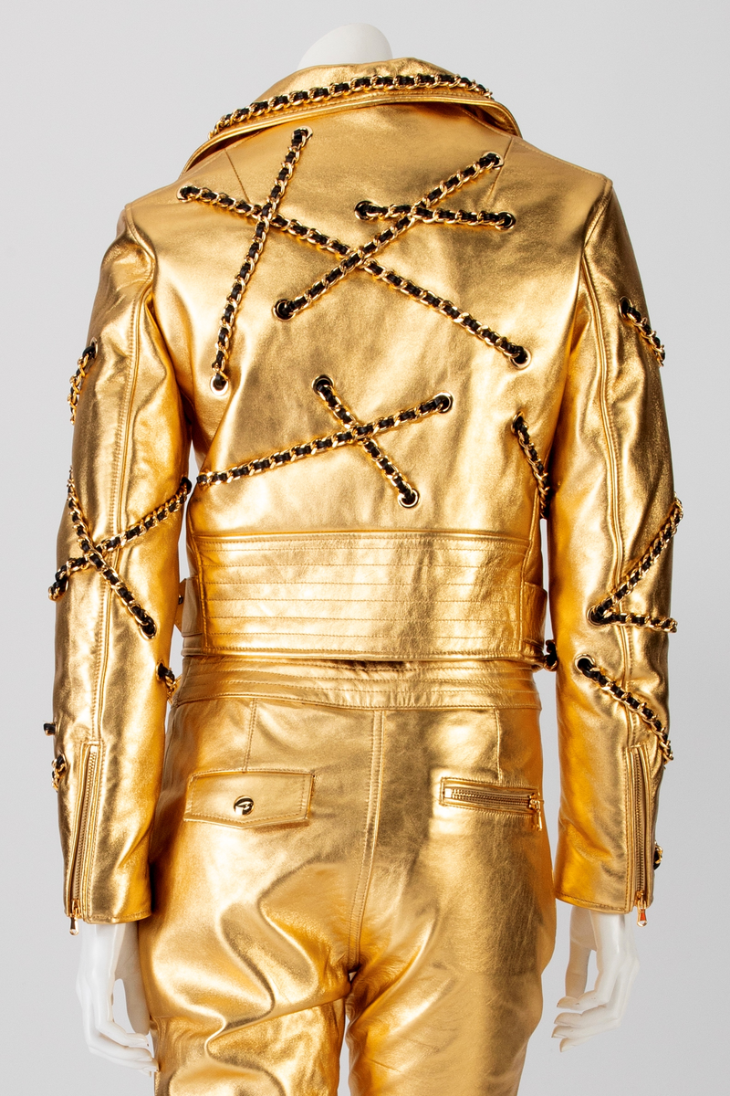 Kort skinnjakke med passepoilerte lommer med gullfarget glidelås og liten lomme med klaff, krage og slag. Lukkes med glidelås midt foran. Tosøms erme med smalt glidelås ved håndleddet. Jakken er dekorert med gullfarget kjetting med skinnsnor smettet inn, tredd gjennom gullfargede maljer på plagget. Dekoren er på krage, forstykke, ermer og bakstykke. Belte med gullfarget spenne i jakkens nederkant.