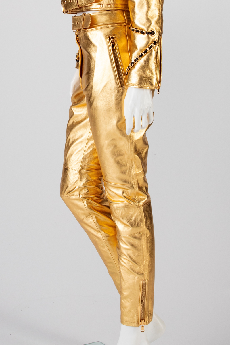 Skinnbukse, jeans-modell, med passepoilerte stikklommer med gullforgylt metallglidelås. På høyre side, nær linningen, en urlomme med klaff og gulltrykknapp. Gylf midt foran med gullforgylt glidelås og trykknapp i linningen. En passepoilert baklomme med gullforgylt glidelås på høyre side. På venstre side er lommen lik urlommen foran. Linningen har fem stikninger. Beltet kommer ut av linningen ved sidesømmen, Beltet har gullforgylt metallspenne og maljer. Formet over kneet. Gullforgylt glidelås med passepoiler ved fotvidden.