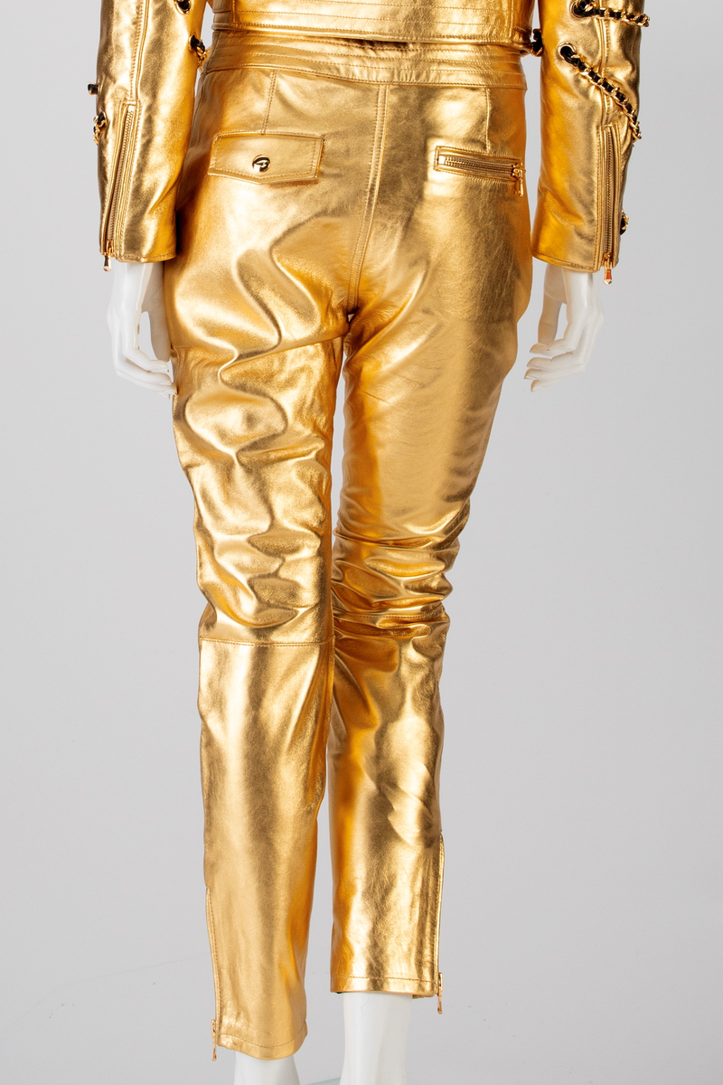 Skinnbukse, jeans-modell, med passepoilerte stikklommer med gullforgylt metallglidelås. På høyre side, nær linningen, en urlomme med klaff og gulltrykknapp. Gylf midt foran med gullforgylt glidelås og trykknapp i linningen. En passepoilert baklomme med gullforgylt glidelås på høyre side. På venstre side er lommen lik urlommen foran. Linningen har fem stikninger. Beltet kommer ut av linningen ved sidesømmen, Beltet har gullforgylt metallspenne og maljer. Formet over kneet. Gullforgylt glidelås med passepoiler ved fotvidden.