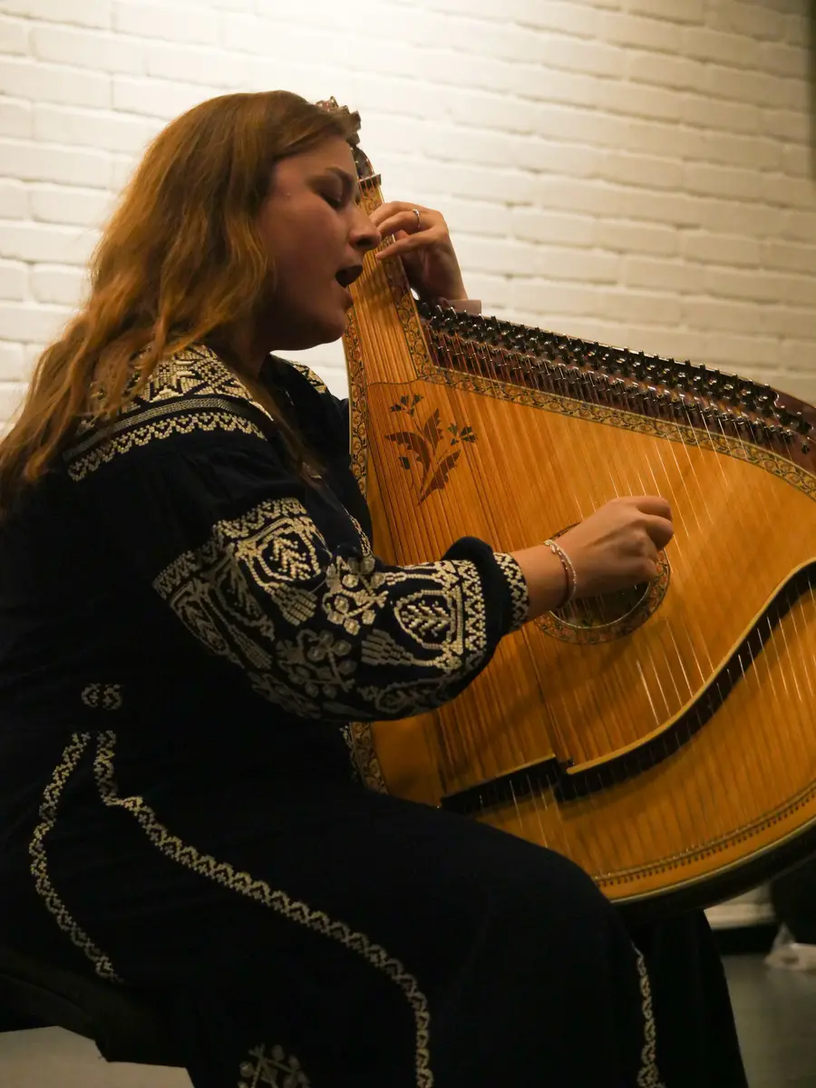 En kvinne med langt hår spiller på et strengeinstrument med mange strenger. Hun har på seg sorte klær med hvit dekor på. En hvit murvegg i bakgrunn.