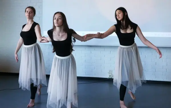 Tre jenter i sorte ballettdrakter og lange, hvite tyllskjørt holder hverandre i armene i dansepositur.