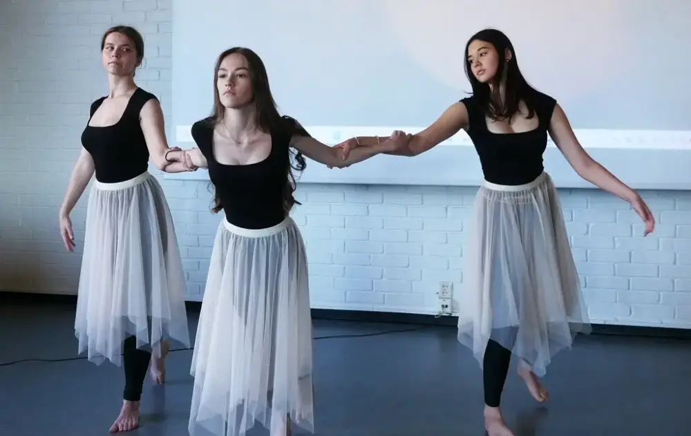 Tre jenter i sorte ballettdrakter og lange, hvite tyllskjørt holder hverandre i armene i dansepositur.