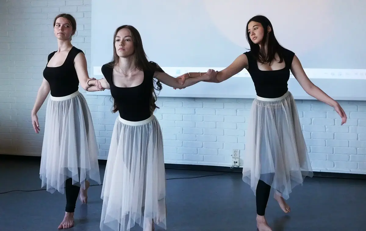 Tre jenter i sorte ballettdrakter og lange, hvite tyllskjørt holder hverandre i armene i dansepositur.