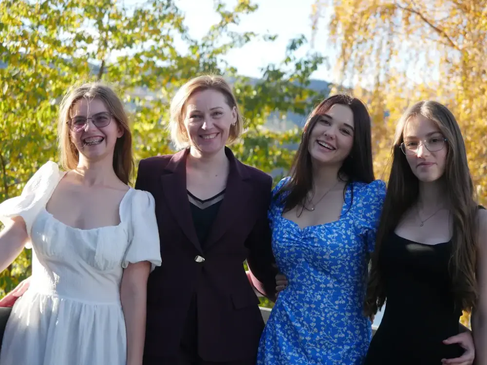 Fire smilende kvinner i kjoler står utendørs med trær i bakgrunn.