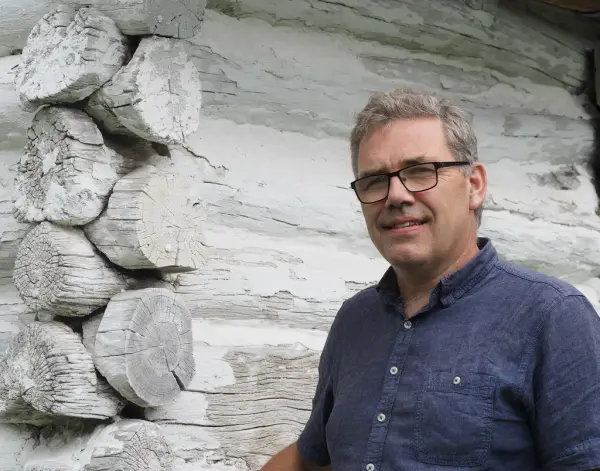 En mann med briller og blå skjorte står foran en hvitkalket tømmervegg.