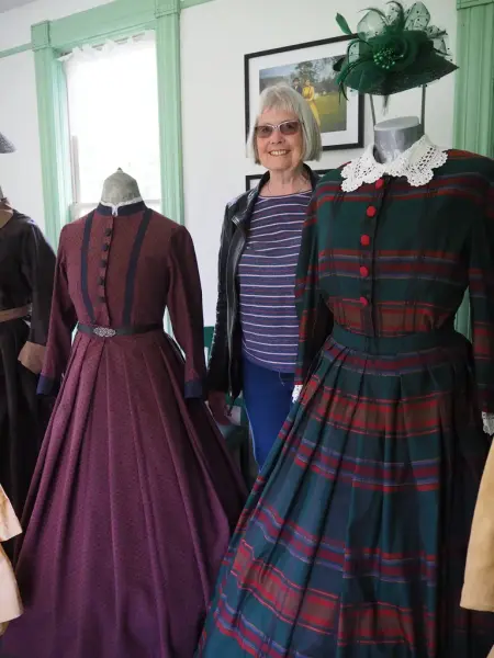 En kvinne står mellom to byster med historiske kostymer på. I bakgrunn et interiør i hvit og lysegrønt.