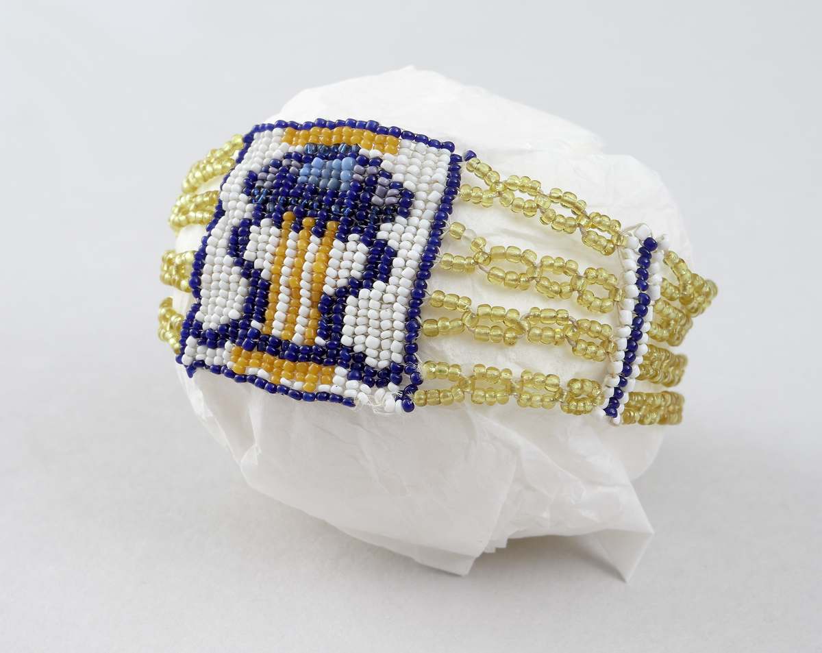 Armband av olikfärgade pärlor i gula, blåa och vita toner. Pärlorna är uppträdda på fyra parallella trådar, gemensamt infästade i metallåsen (möjligen av silver, stämplad NR på baksidan). Armbandets motiv finns mittemot låset och är ett lyrformat pärlbroderi i olika färger.
Inskrivet i huvudkatalog 1898 -1900.