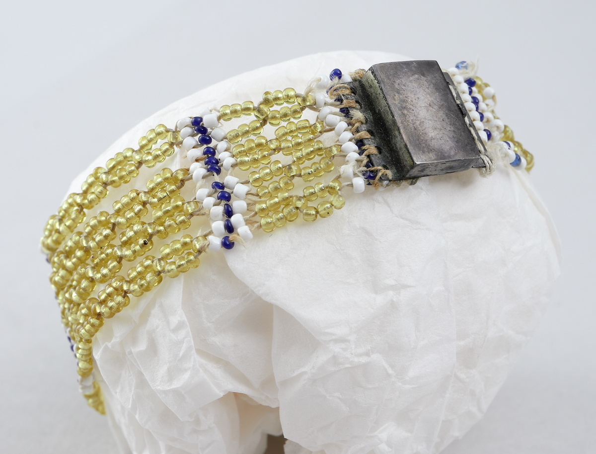 Armband av olikfärgade pärlor i gula, blåa och vita toner. Pärlorna är uppträdda på fyra parallella trådar, gemensamt infästade i metallåsen (möjligen av silver, stämplad NR på baksidan). Armbandets motiv finns mittemot låset och är ett lyrformat pärlbroderi i olika färger.
Inskrivet i huvudkatalog 1898 -1900.