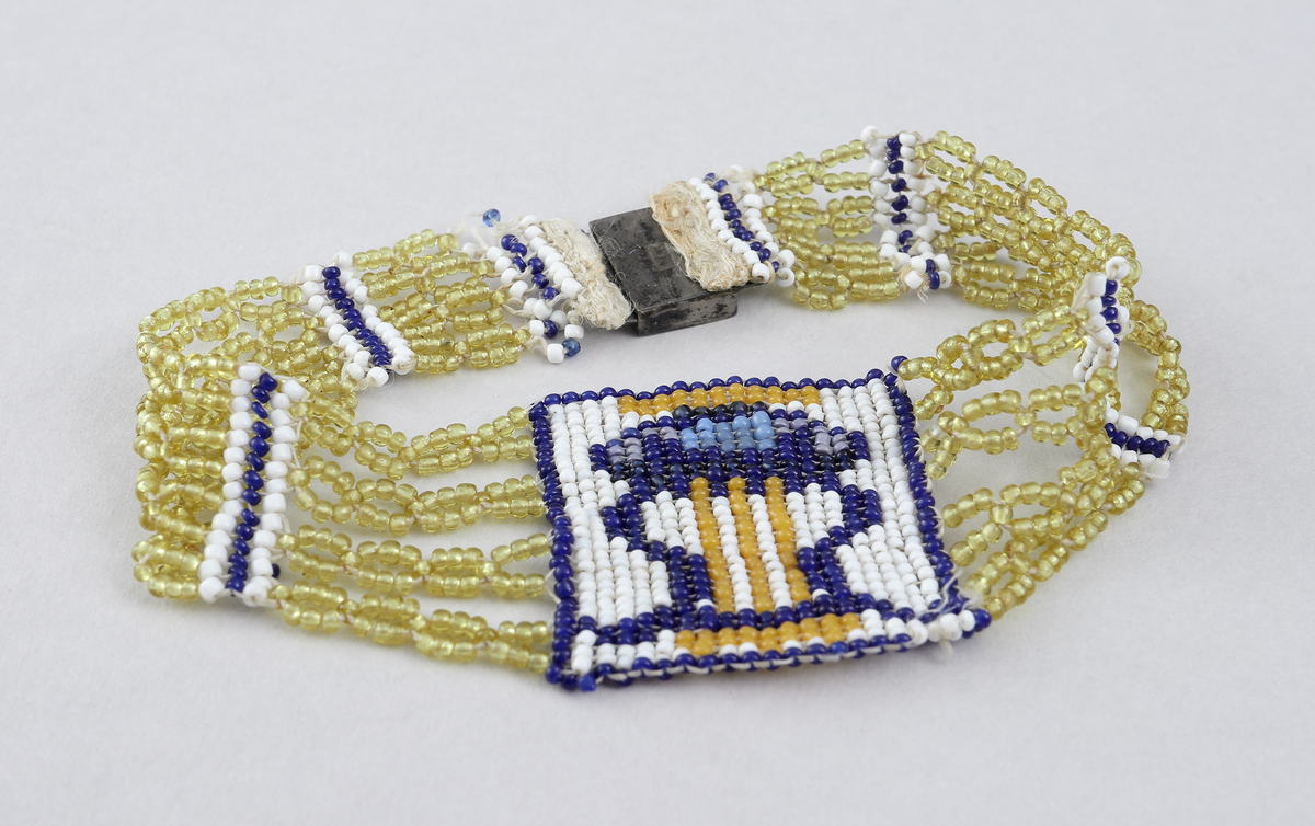 Armband av olikfärgade pärlor i gula, blåa och vita toner. Pärlorna är uppträdda på fyra parallella trådar, gemensamt infästade i metallåsen (möjligen av silver, stämplad NR på baksidan). Armbandets motiv finns mittemot låset och är ett lyrformat pärlbroderi i olika färger.
Inskrivet i huvudkatalog 1898 -1900.