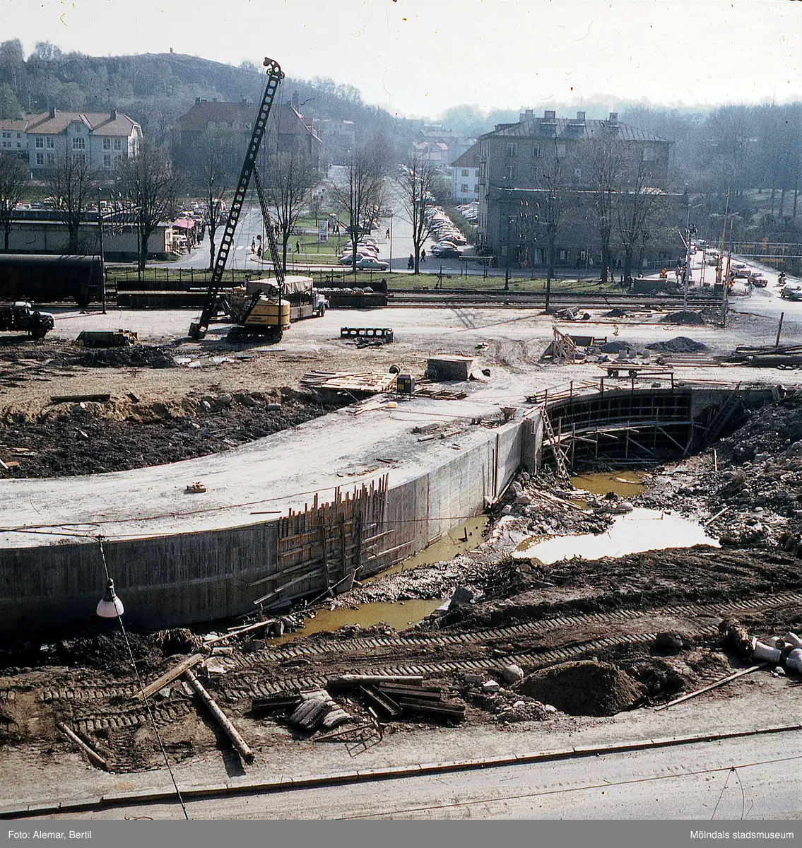 Muddring av Mölndalsån inför planerad byggnation av Mölndals bro. Fotot ser ut att vara taget när det är vår på grund av det gröna gräset, men att det inte finns några löv på träden. Tid: Omkring 1974. I bakgrunden syns Träskolan, Nya Torget och stadsdelen Trädgården. Kvarnbyskolan syns till vänster i bild.
Bilar står parkerade i långa rader.