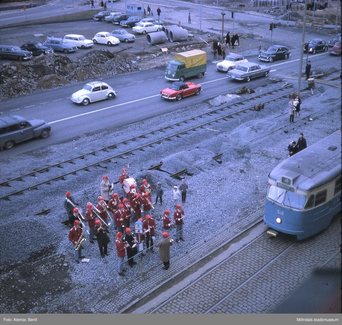 Muddring av Mölndalsån i bakgrunden inför planerad byggnation av Mölndals bro. 
Tid: Cirka slutet av 1960-talet. 
Bilden visar stadstrafik samt anläggningsarbete. I mitten av bilden skymtar den nya spårvagnsrälsen som håller på att anläggas. En liten musikkår som bär tomteluvor, står och spelar julsånger nära spårvagnshållplatsen. Tre barn, en flicka och två pojkar, står i närheten av musikkåren och tittar på och lyssnar.
På spårvagnen för linje fyra, står det att den går mot Kungssten. De två äldre parallella spårvagnsspåren som går mellan Göteborg och Mölndals bro, belagd med kullersten, syns till höger i förgrunden.