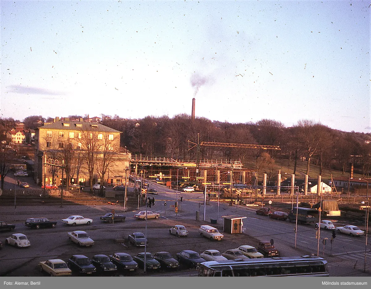 Långt framskriden byggnation av Mölndals bro. I bakgrunden, bakom pelarna skymtar även vita, låga byggnader som är växthus och ligger vid foten av parken som hör till Villa Korndal. 
Tid: Cirka början av 1970-talet. 
Bilden visar stadstrafik samt anläggningsarbete. 
I bakgrunden skymtar den stora skorstenen som finns vid den här tiden på Papyrus område. I förgrunden syns flertalet personbilar parkerade.