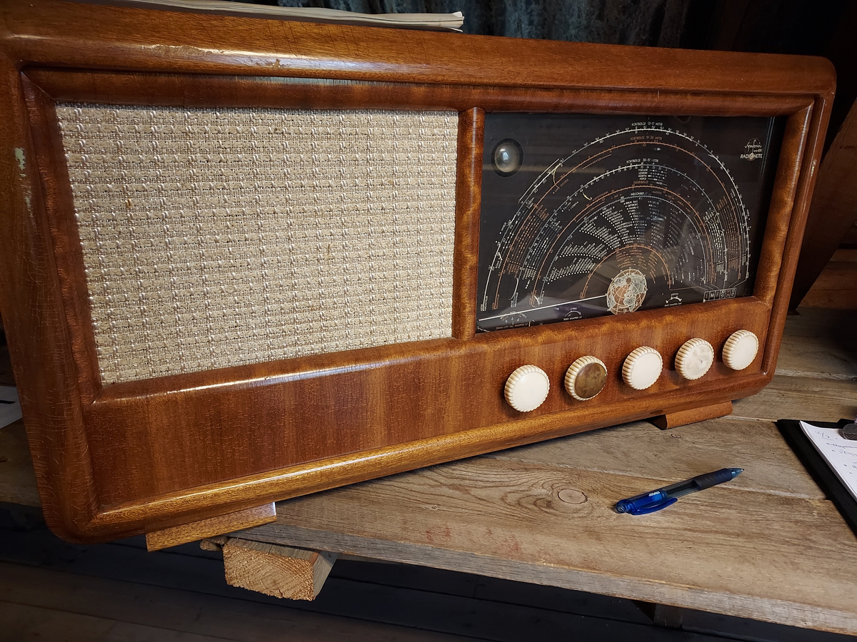 Radio. Lakkert treverk. Teak. Beige tekstil foran høytaler. Buet søkerskive i hvit og rødt. Jordklode på midten nederst. Logo på hver side øverst på søkerskive. Trolløye på venstre side. Sort bakgrunn, glass foran. 4 hvite søkerknotter. En mangler. Brun perforert pappskive på baksiden med diverse informasjon og papiretiketter. Radioen er elektrisk. Sort ledning. Sort støpsel. Radioen står på 4 lave treklosser.
