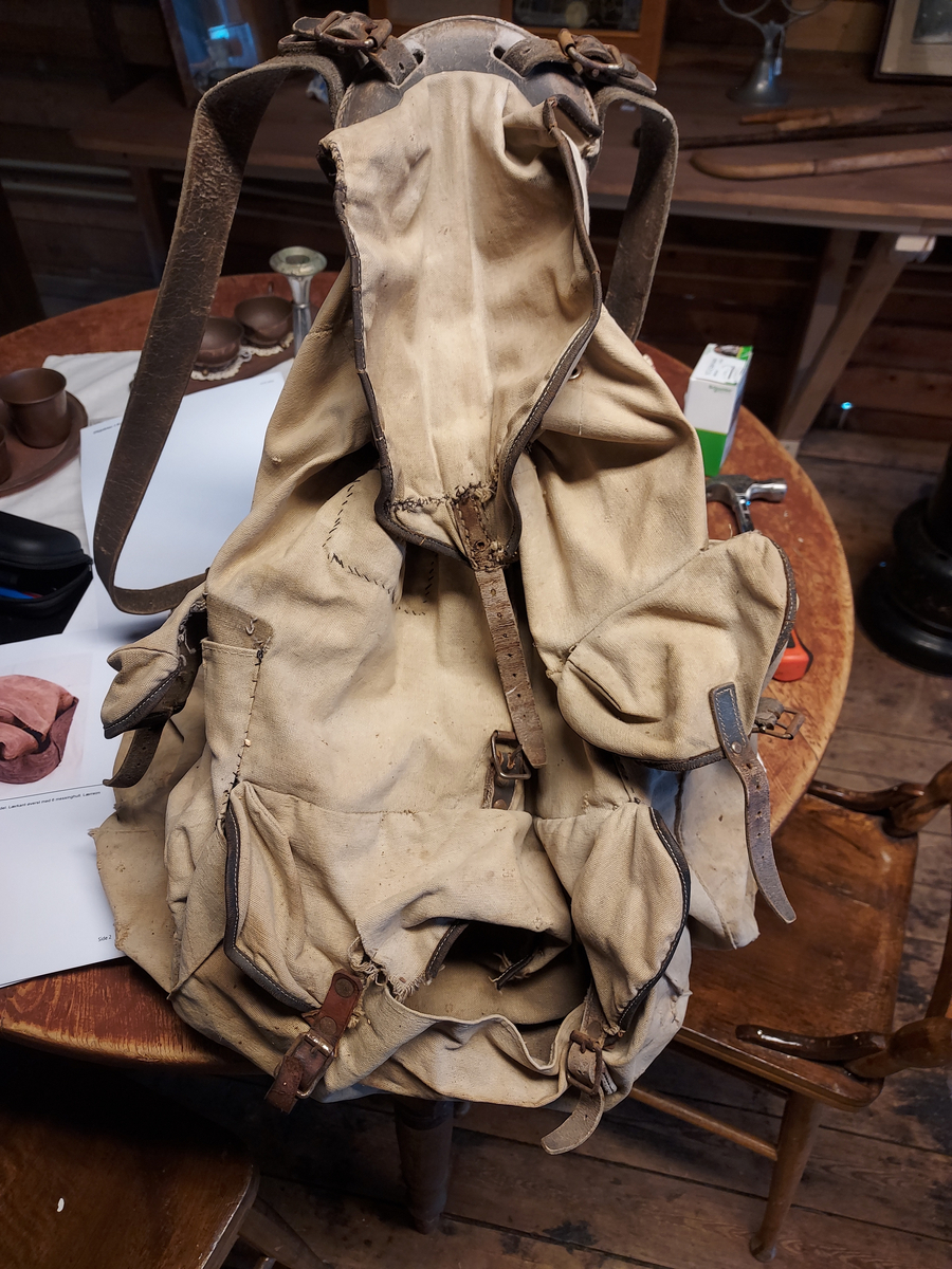 Ryggsekk. Beige tekstil. Meis i metall. Lærreimer til forsterkning og innstramming. Mye reparert med søm. To sidelommer og en midt på ved bunnen foran. Lukkes med klaff og innsnøring.