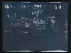 Ett stort sällskap fikar vid ett bord, utomhus bland tallar. Fotografens anteckning på negativet 