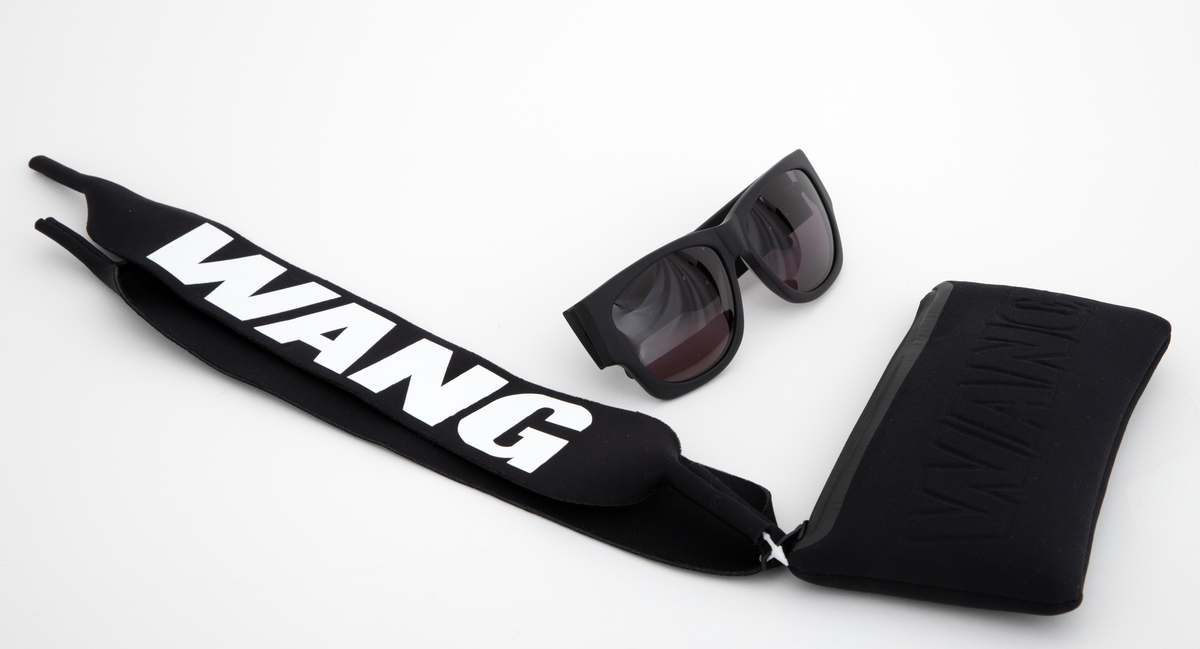 Relativt store, men lette solbriller av matt, svart plast. Mens innfatningen er i hardplast, virker stengene gummiert i overflaten. Stengene er brede og gjennombrudt i fremre del, og utfylt med WANG i store bokstaver. Plastglassene er mørke og gråfiolette. Brilleetuiet er av tynt, neoprenlignende materiale, med gummiert plastglidelås. Den ene siden har bokstavene WANG trykket ned i materialet, mens den andre siden har teksten ALEXANDER WANG x H&M i forhøyet gummitrykk. Brillesnorer, to stykker av neopren, til å tre innpå stengene, den ene pålimt navnet WANG i hvite plastbokstaver, den andre WANG i svarte bokstaver.