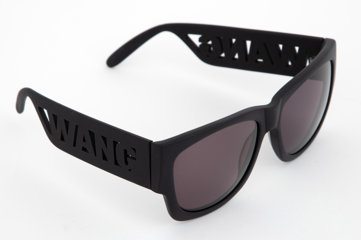 Relativt store, men lette solbriller av matt, svart plast. Mens innfatningen er i hardplast, virker stengene gummiert i overflaten. Stengene er brede og gjennombrudt i fremre del, og utfylt med WANG i store bokstaver. Plastglassene er mørke og gråfiolette. Brilleetuiet er av tynt, neoprenlignende materiale, med gummiert plastglidelås. Den ene siden har bokstavene WANG trykket ned i materialet, mens den andre siden har teksten ALEXANDER WANG x H&M i forhøyet gummitrykk. Brillesnorer, to stykker av neopren, til å tre innpå stengene, den ene pålimt navnet WANG i hvite plastbokstaver, den andre WANG i svarte bokstaver.
