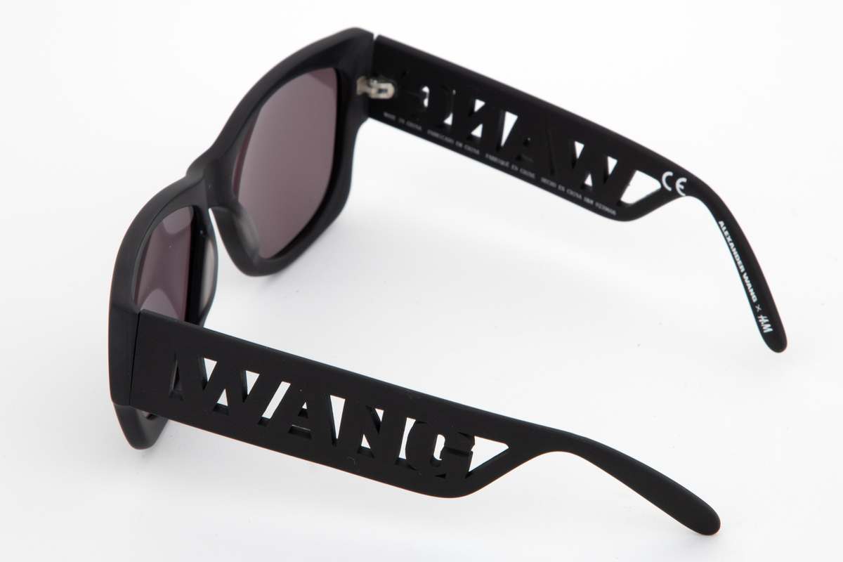 Relativt store, men lette solbriller av matt, svart plast. Mens innfatningen er i hardplast, virker stengene gummiert i overflaten. Stengene er brede og gjennombrudt i fremre del, og utfylt med WANG i store bokstaver. Plastglassene er mørke og gråfiolette. Brilleetuiet er av tynt, neoprenlignende materiale, med gummiert plastglidelås. Den ene siden har bokstavene WANG trykket ned i materialet, mens den andre siden har teksten ALEXANDER WANG x H&M i forhøyet gummitrykk. Brillesnorer, to stykker av neopren, til å tre innpå stengene, den ene pålimt navnet WANG i hvite plastbokstaver, den andre WANG i svarte bokstaver.