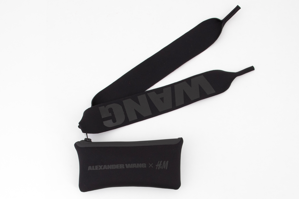 Relativt store, men lette solbriller av matt, svart plast. Mens innfatningen er i hardplast, virker stengene gummiert i overflaten. Stengene er brede og gjennombrudt i fremre del, og utfylt med WANG i store bokstaver. Plastglassene er mørke og gråfiolette. Brilleetuiet er av tynt, neoprenlignende materiale, med gummiert plastglidelås. Den ene siden har bokstavene WANG trykket ned i materialet, mens den andre siden har teksten ALEXANDER WANG x H&M i forhøyet gummitrykk. Brillesnorer, to stykker av neopren, til å tre innpå stengene, den ene pålimt navnet WANG i hvite plastbokstaver, den andre WANG i svarte bokstaver.