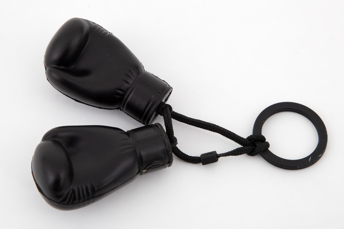 To små, myke boksehansker som henger i en svart snor fra en splittet ring av hardplast. Hanskene viser en "knyttet" hånd, og på yttersiden vises de sammentrukne bokstavene AW.