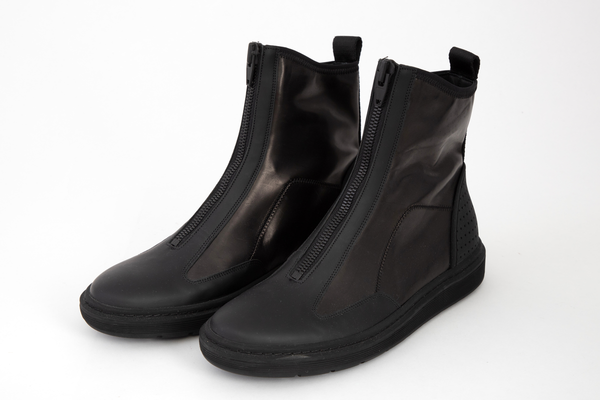 Ankelhøye herrestøvler i svart skinn med kraftig, svart glidelås midt foran. På baksiden påsydd bånd med hempe øverst og innvevd logo: WANG. Kraftig profilert såle med bokstavene WA etter hverandre.