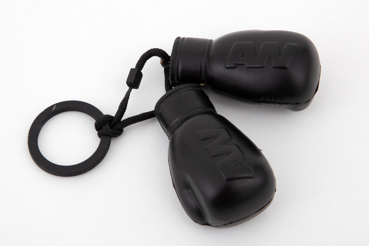 To små, myke boksehansker som henger i en svart snor fra en splittet ring av hardplast. Hanskene viser en "knyttet" hånd, og på yttersiden vises de sammentrukne bokstavene AW.