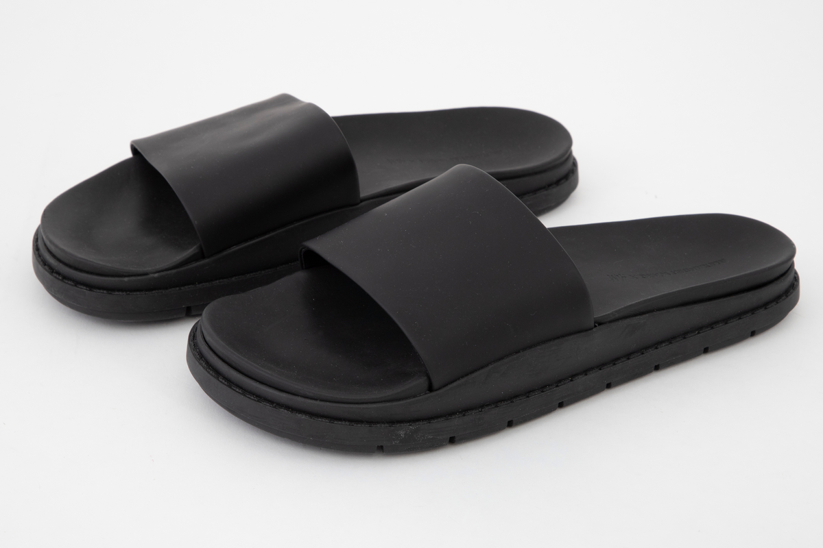 Svarte sandaler med matt og "gummiert" overflate, merket som lær. Kraftig såle med bred "stropp" over fotbladet. Åpen tå, ingen hælkappe eller hælstropp. Innsiden av "stroppen", overlæret, er fôret med svart tekstil. Innsiden av sålen er ergonomisk støpt, med oppbygning under gelenken og opphøyde kanter rundt fotsengen. Det er samme overflate på innsiden av sålen, dvs. en matt, gummiaktig overflate, antagelig vannavstøtende. Sålen gir inntrykk av å være randsydd, men er antagelig støpt. Oppgis å være syntetmateriale. Undersiden er dypt mønstret, med parallelle rekker med bokstavene AWAWAWAW, osv.