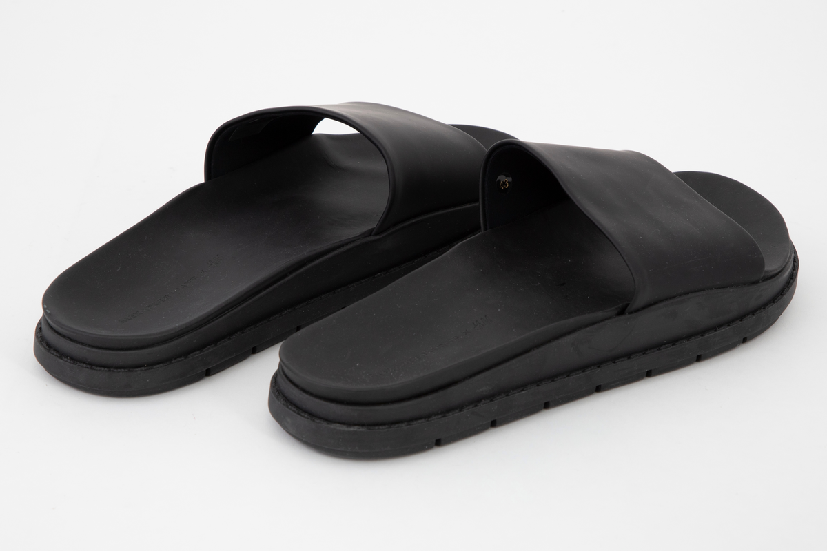 Svarte sandaler med matt og "gummiert" overflate, merket som lær. Kraftig såle med bred "stropp" over fotbladet. Åpen tå, ingen hælkappe eller hælstropp. Innsiden av "stroppen", overlæret, er fôret med svart tekstil. Innsiden av sålen er ergonomisk støpt, med oppbygning under gelenken og opphøyde kanter rundt fotsengen. Det er samme overflate på innsiden av sålen, dvs. en matt, gummiaktig overflate, antagelig vannavstøtende. Sålen gir inntrykk av å være randsydd, men er antagelig støpt. Oppgis å være syntetmateriale. Undersiden er dypt mønstret, med parallelle rekker med bokstavene AWAWAWAW, osv.