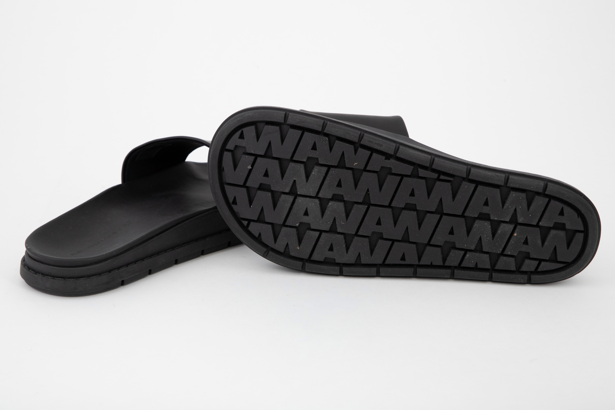 Svarte sandaler med matt og "gummiert" overflate, merket som lær. Kraftig såle med bred "stropp" over fotbladet. Åpen tå, ingen hælkappe eller hælstropp. Innsiden av "stroppen", overlæret, er fôret med svart tekstil. Innsiden av sålen er ergonomisk støpt, med oppbygning under gelenken og opphøyde kanter rundt fotsengen. Det er samme overflate på innsiden av sålen, dvs. en matt, gummiaktig overflate, antagelig vannavstøtende. Sålen gir inntrykk av å være randsydd, men er antagelig støpt. Oppgis å være syntetmateriale. Undersiden er dypt mønstret, med parallelle rekker med bokstavene AWAWAWAW, osv.