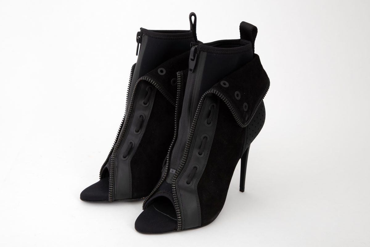 Svart, ankelhøy skolett med åpen, spiss tå, høy, syltynn stiletthæl, tynn, men laminert såle og glidelåslukning midt foran. Konstruksjonen består av to lag materiale: det innerste av neopren, lukket med vanntett, sveiset glidelås, det ytre av tynt semsket skinn, forsterket med tekstil på baksiden. Det innerste laget kan dras på ved hjelp av en bred stropp midt bak. Det ytre skallet er høyere enn det indre og kan brettes ned og kneppes med trykknapp midt bak. Det er dekorativt kantet med kraftig metallglidelås i overkant og ned langs fronten. Fronten er i tillegg forsterket med et ca. 3 cm bredt bånd av plastlignende materiale, utstyrt med 10 maljer på hver side, de 6 nederste itredd kraftig skolisse. Hælkappen er forsterket med gummi, med ruglete struktur og tettstilte hull som dekor. Stiletthælen og sålen er av gummiert lær. Også innersålen skal være av skinn.