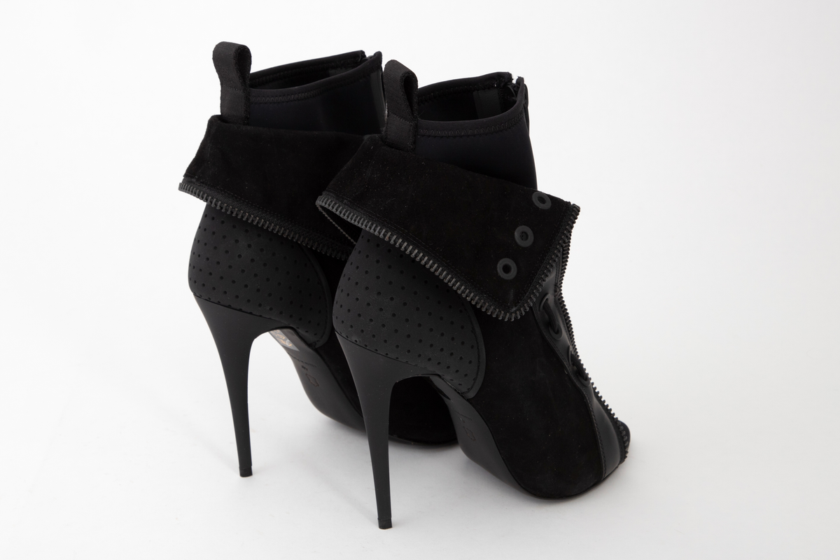 Svart, ankelhøy skolett med åpen, spiss tå, høy, syltynn stiletthæl, tynn, men laminert såle og glidelåslukning midt foran. Konstruksjonen består av to lag materiale: det innerste av neopren, lukket med vanntett, sveiset glidelås, det ytre av tynt semsket skinn, forsterket med tekstil på baksiden. Det innerste laget kan dras på ved hjelp av en bred stropp midt bak. Det ytre skallet er høyere enn det indre og kan brettes ned og kneppes med trykknapp midt bak. Det er dekorativt kantet med kraftig metallglidelås i overkant og ned langs fronten. Fronten er i tillegg forsterket med et ca. 3 cm bredt bånd av plastlignende materiale, utstyrt med 10 maljer på hver side, de 6 nederste itredd kraftig skolisse. Hælkappen er forsterket med gummi, med ruglete struktur og tettstilte hull som dekor. Stiletthælen og sålen er av gummiert lær. Også innersålen skal være av skinn.