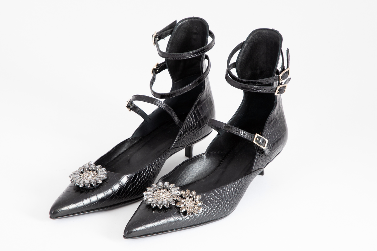 Svært spisse, dypt ringede lærsko med svært lave stiletthæler. Hælkappen er høy i bakkant, dvs. over ankelhøyde, og skoen holdes på plass med to tynne ankelstropper og en vristrem. Ekstra forsterkning over sømmen midt bak, tjener som smygstol for ankelremmene. Alle remmene kan reguleres og lukkes med messingfarget spenne. Innvendig fôret med sort skinn, og beskjeden polstring under hælen. Flikk og såle er også sorte. Læret er preget med krokodille- eller alligatormønster. Skoene er ellers prydet med sammenlenkede, klare og grå blomster av glasstener, såkalt strass.