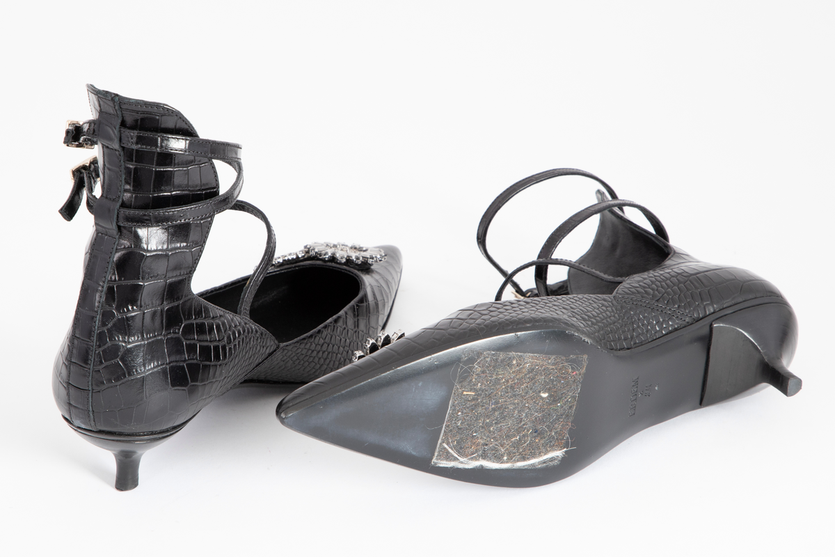 Svært spisse, dypt ringede lærsko med svært lave stiletthæler. Hælkappen er høy i bakkant, dvs. over ankelhøyde, og skoen holdes på plass med to tynne ankelstropper og en vristrem. Ekstra forsterkning over sømmen midt bak, tjener som smygstol for ankelremmene. Alle remmene kan reguleres og lukkes med messingfarget spenne. Innvendig fôret med sort skinn, og beskjeden polstring under hælen. Flikk og såle er også sorte. Læret er preget med krokodille- eller alligatormønster. Skoene er ellers prydet med sammenlenkede, klare og grå blomster av glasstener, såkalt strass.