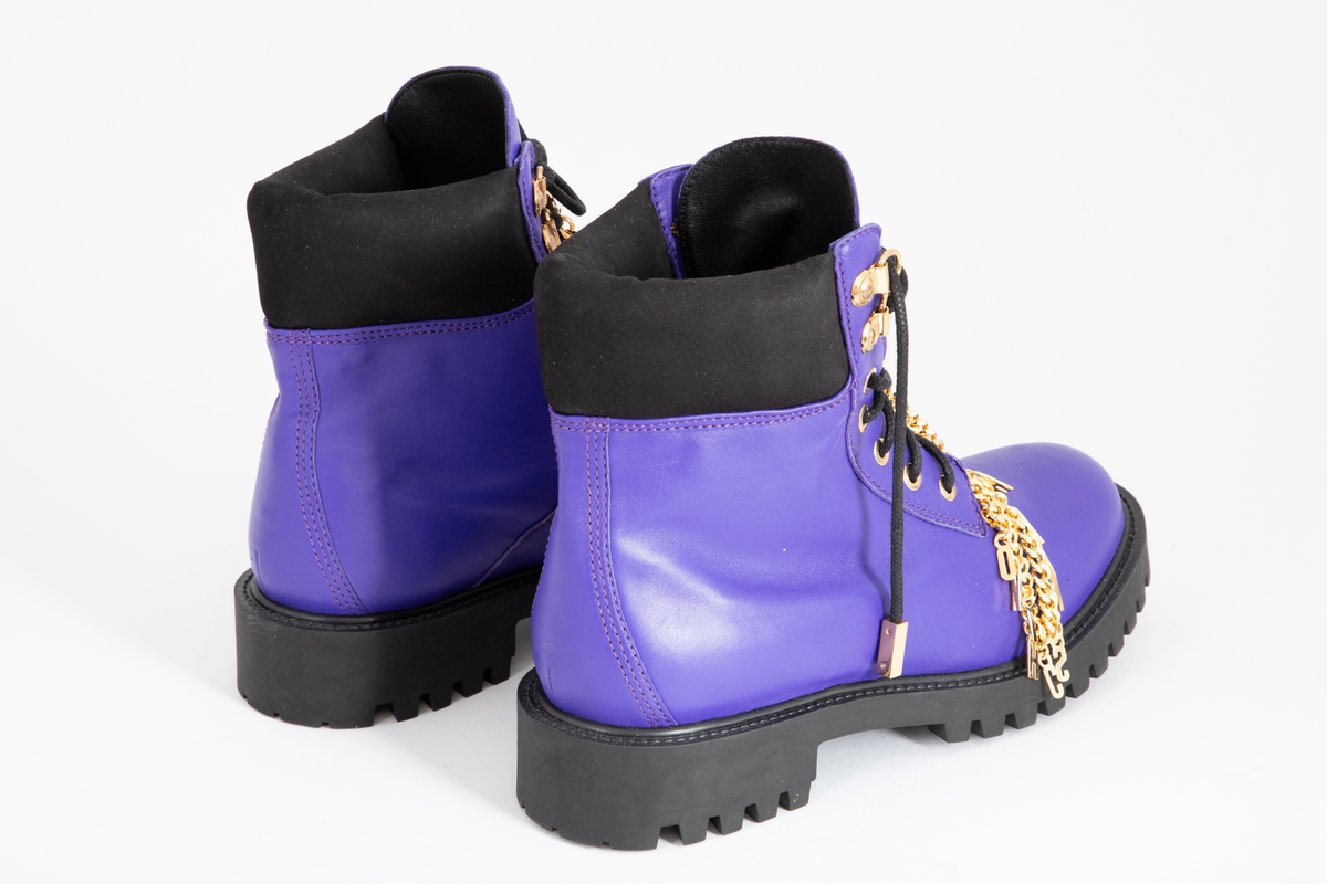 Damestøvler i blåfiolett skinn med kraftig gummisåle. Avrundet foran, med sju messingmaljer på hver side og svart, rund og kraftig skoreim. Messinglenke med MOSCHINO-logo hengende nedover skoen.