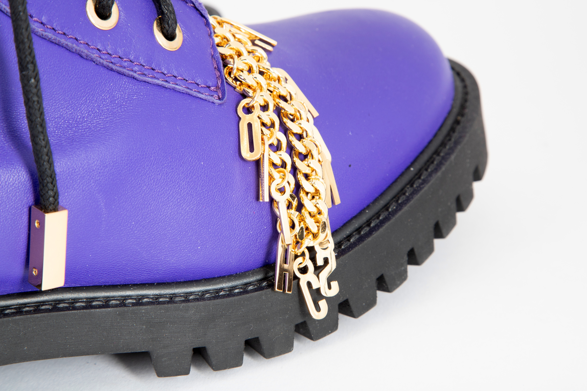 Damestøvler i blåfiolett skinn med kraftig gummisåle. Avrundet foran, med sju messingmaljer på hver side og svart, rund og kraftig skoreim. Messinglenke med MOSCHINO-logo hengende nedover skoen.