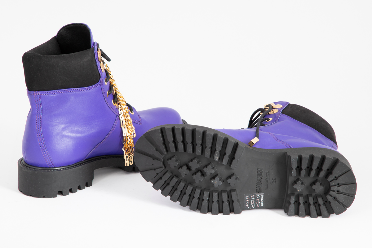 Damestøvler i blåfiolett skinn med kraftig gummisåle. Avrundet foran, med sju messingmaljer på hver side og svart, rund og kraftig skoreim. Messinglenke med MOSCHINO-logo hengende nedover skoen.