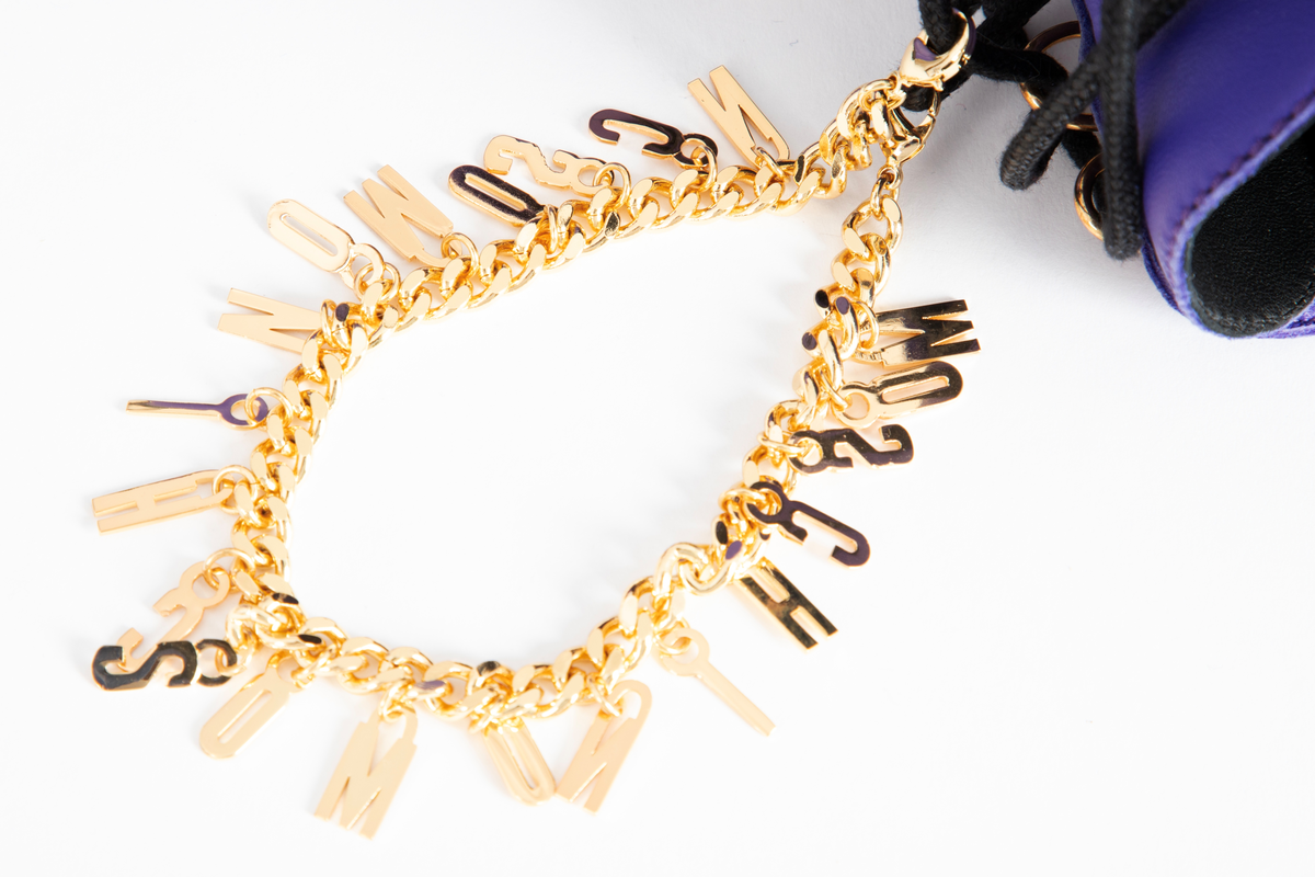 Damestøvler i blåfiolett skinn med kraftig gummisåle. Avrundet foran, med sju messingmaljer på hver side og svart, rund og kraftig skoreim. Messinglenke med MOSCHINO-logo hengende nedover skoen.