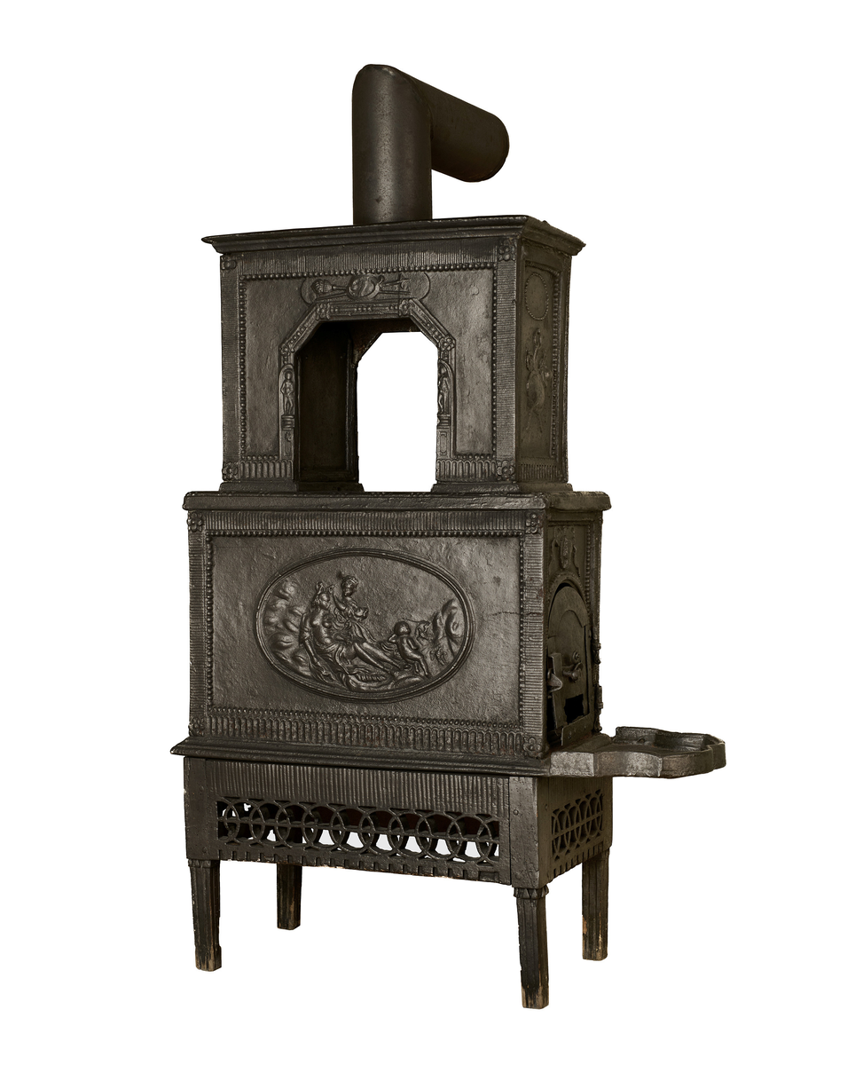 Ovn i to etasjer, utført i støpejern. Den bæres av en trebase med gjennombrutte sirkelornamenter, som hviler på fire kanelerte ben. Samtlige sider er innrammet med perlerad, blomsterornamenter og kanelyrer. Frontpanelet på nedre del viser en tondo med Venus i toalettet, flankert av en tjenerinne og Amor, fremstilt i lavt relieff. Det høyre sidefeltet har ovnsluke og askebrett. Frontpanelet i andre etasje er gjennombrutt med knekkede hjørner, og gjenopptar dekorprogrammet fra nedre del - inkludert spolier og klassiserende figurfremstillinger. Fremskutt, profilert toppstykke, som avsluttes med et sylinderformet ovnsrør med knekk som forbinder ovnen til brannmuren.