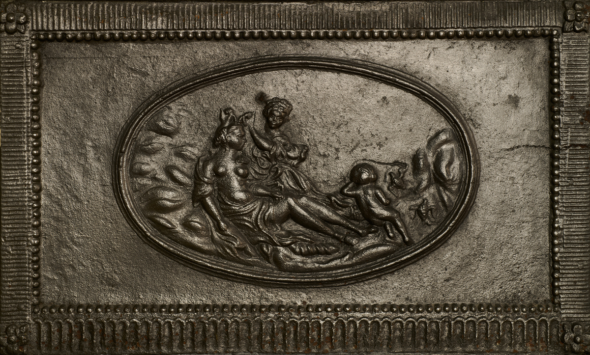 Ovn i to etasjer, utført i støpejern. Den bæres av en trebase med gjennombrutte sirkelornamenter, som hviler på fire kanelerte ben. Samtlige sider er innrammet med perlerad, blomsterornamenter og kanelyrer. Frontpanelet på nedre del viser en tondo med Venus i toalettet, flankert av en tjenerinne og Amor, fremstilt i lavt relieff. Det høyre sidefeltet har ovnsluke og askebrett. Frontpanelet i andre etasje er gjennombrutt med knekkede hjørner, og gjenopptar dekorprogrammet fra nedre del - inkludert spolier og klassiserende figurfremstillinger. Fremskutt, profilert toppstykke, som avsluttes med et sylinderformet ovnsrør med knekk som forbinder ovnen til brannmuren.