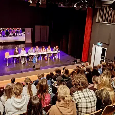 Bildet er tatt fra oppe i en tribune og viser publikum som ser mot scenen med et panel på syv personer som sitter bak et langt bord.