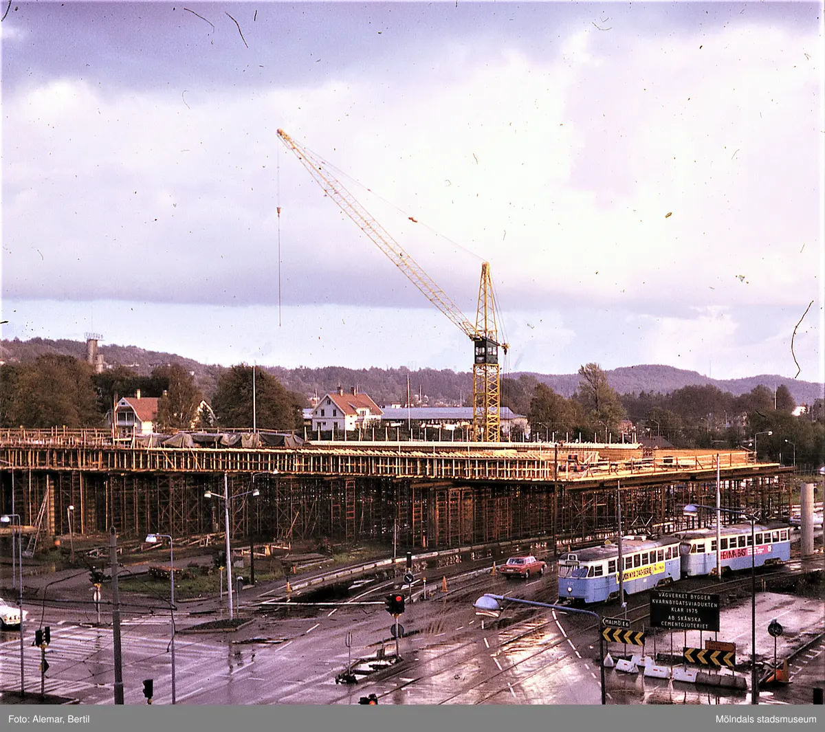Långt framskriden byggnation av Mölndals bro, ser ut att vara taget under någon gång i slutet på våren eller i början av sommarmånaderna juni till augusti.
Tid: Cirka början av 1970-talet. 
Bilden visar stadstrafik samt anläggningsarbete. 
I bakgrunden till vänster skymtar tornet med företagsnamnet Papyrus.  
I förgrunden syns flertalet personbilar som passerar.
Längst ned till höger skymtar spårgvagnsspåren och del av den gamla vändslingan för spårvagnen, innan den blir överbyggd. Den första spårvagnen, som drar en vagn bakom sig, har på sin vänstra sida en gul, rektangulär reklamaffisch uppsatt, med texten: "VOLVOS NYA 240-SERIE". Längre ned till höger skymtar en svart vägtrafikskylt med vit skrift. Skylten har följande text: "VÄGVERKET BYGGER KVANRBYGATSVIADUKTEN KLART 1975 AB SKÅNSKA CEMENTGJUTERIET".