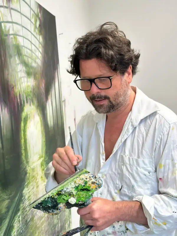 Mann maler et bilde med grønne farger. Han er ikledd hvit skjorte og har på seg briller med sort innfatning. 