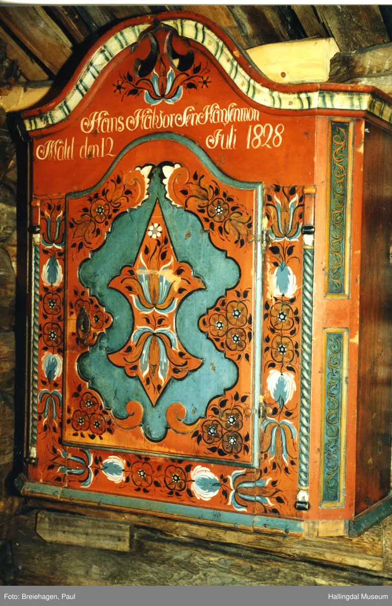 Hengeskap H. m/gavl og kantlist: 104,6- H.skap: 87,2- Br. 85,4- D. 36,1 - H.dør: 68,5- Br.dør: 48,2. Gran, snekret,malt. Delene holdt sammen v.hj.av tapping og trenagler. Gavlen buet oppover. Kronlisten, hulkil med vulst på hver side. I toppen på gavlen gj.gående utskjæring. 2 C-er med ryggen mot hverandre. Skapet rektangulært med avkuttede hjørner i fronten.