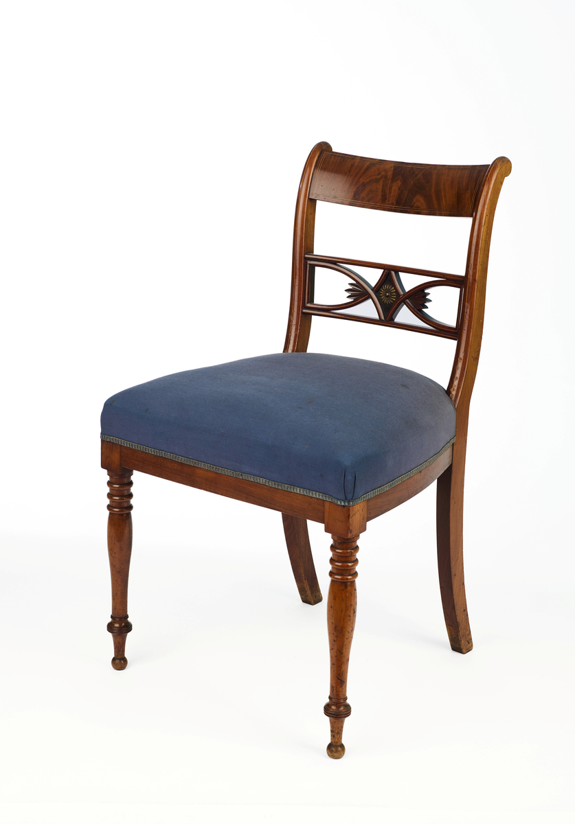 Stol av finért mahogni, som hviler på fire ben. Åpen rygg med glatt buet toppstykke med intarsia i lyst tre. Gjennombrutt tverrspross med halvsirkelformede ornamenter, hvor det ovale midtpartiet er markert med en riflet metallrosett. Kurvede akterstaver glir over i bakbenene, mens forbenene er dreide. Trapesformet sete med sekundært blåfarget trekk i ull. Mykt buet sarg, hvis sider er svakt trukket inn under setet.
