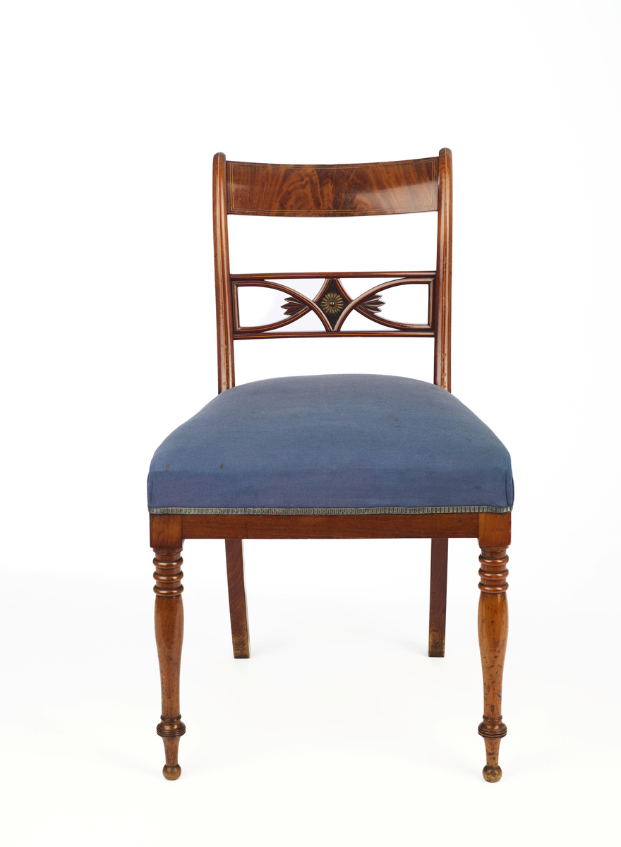 Stol av finért mahogni, som hviler på fire ben. Åpen rygg med glatt buet toppstykke med intarsia i lyst tre. Gjennombrutt tverrspross med halvsirkelformede ornamenter, hvor det ovale midtpartiet er markert med en riflet metallrosett. Kurvede akterstaver glir over i bakbenene, mens forbenene er dreide. Trapesformet sete med sekundært blåfarget trekk i ull. Mykt buet sarg, hvis sider er svakt trukket inn under setet.
