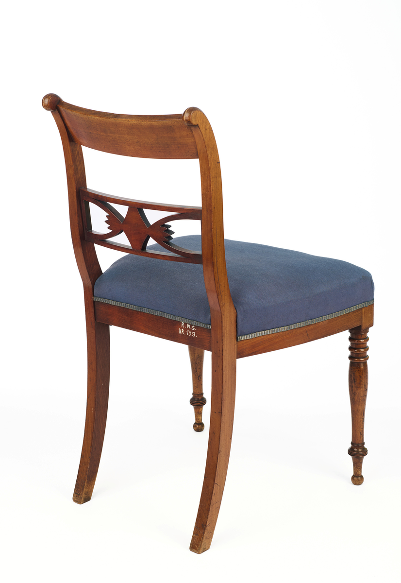 Stol av finért mahogni, som hviler på fire ben. Åpen rygg med glatt buet toppstykke med intarsia i lyst tre. Gjennombrutt tverrspross med halvsirkelformede ornamenter, hvor det ovale midtpartiet er markert med en riflet metallrosett. Kurvede akterstaver glir over i bakbenene, mens forbenene er dreide. Trapesformet sete med sekundært blåfarget trekk i ull. Mykt buet sarg, hvis sider er svakt trukket inn under setet.
