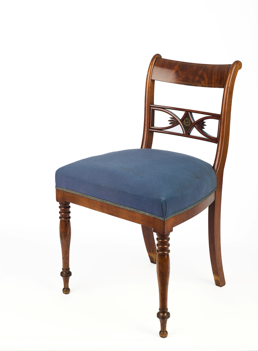 Stol av finért mahogni, som hviler på fire ben. Åpen rygg med glatt buet toppstykke med intarsia i lyst tre. Gjennombrutt tverrspross med halvsirkelformede ornamenter, hvor det ovale midtpartiet er markert med en riflet metallrosett. Kurvede akterstaver glir over i bakbenene, mens forbenene er dreide. Trapesformet sete med sekundært blåfarget trekk i ull. Mykt buet sarg, hvis sider er svakt trukket inn under setet.
