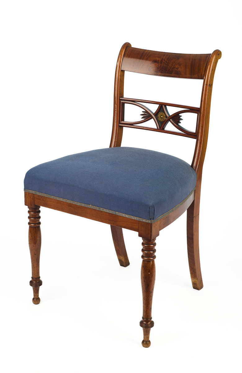 Stol av finért mahogni, som hviler på fire ben. Åpen rygg med glatt buet toppstykke med intarsia i lyst tre. Gjennombrutt tverrspross med halvsirkelformede ornamenter, hvor det ovale midtpartiet er markert med en riflet metallrosett. Kurvede akterstaver glir over i bakbenene, mens forbenene er dreide. Trapesformet sete med sekundært blåfarget trekk i ull. Mykt buet sarg, hvis sider er svakt trukket inn under setet.