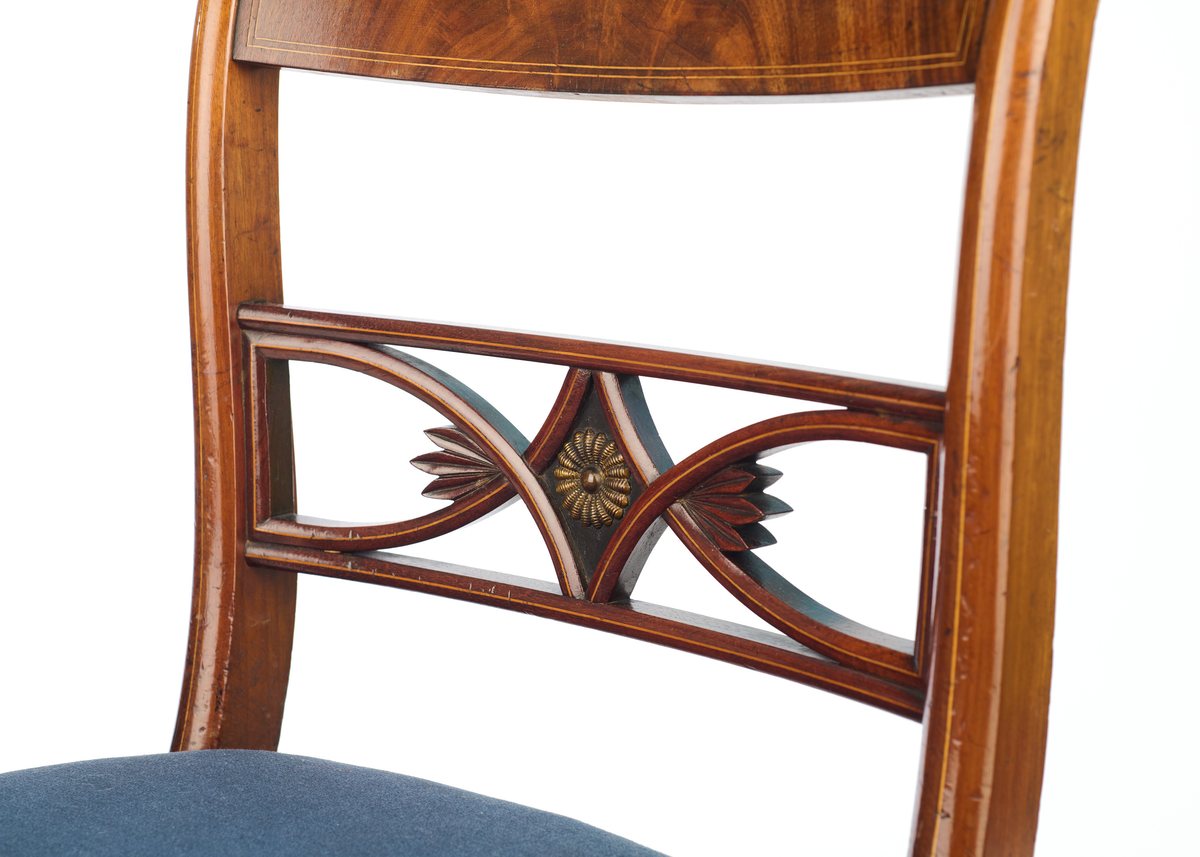 Stol av finért mahogni, som hviler på fire ben. Åpen rygg med glatt buet toppstykke med intarsia i lyst tre. Gjennombrutt tverrspross med halvsirkelformede ornamenter, hvor det ovale midtpartiet er markert med en riflet metallrosett. Kurvede akterstaver glir over i bakbenene, mens forbenene er dreide. Trapesformet sete med sekundært blåfarget trekk i ull. Mykt buet sarg, hvis sider er svakt trukket inn under setet.