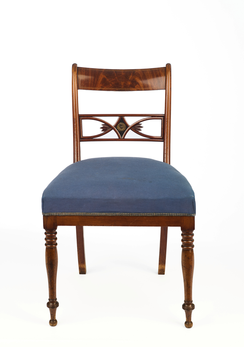 Stol av finért mahogni, som hviler på fire ben. Åpen rygg med glatt buet toppstykke med intarsia i lyst tre. Gjennombrutt tverrspross med halvsirkelformede ornamenter, hvor det ovale midtpartiet er markert med en riflet metallrosett. Kurvede akterstaver glir over i bakbenene, mens forbenene er dreide. Trapesformet sete med sekundært blåfarget trekk i ull. Mykt buet sarg, hvis sider er svakt trukket inn under setet.